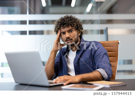 Portrait of a young shocked Indian man sitting at a desk in the office with a laptop, holding his head in frustration and looking at the camera. Portrait of a young shocked Indian man sitting at a desk in the office with a laptop, holding his head in frustration and looking at the camera. 137944413