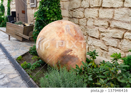 A large, brown, ceramic vase is sitting on the ground next to some plants A large, brown, ceramic vase is sitting on the ground next to some plants 137946416
