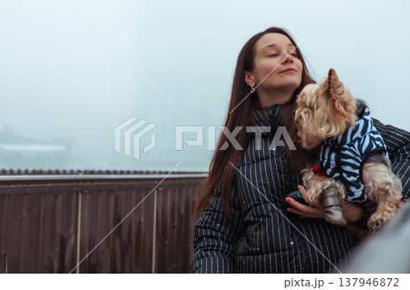 A beautiful young woman in a stylish puffer jacket holding a cute Yorkshire Terrier dog outdoors on a misty winter day. A bond between owner and pet. A beautiful young woman in a stylish puffer jacket holding a cute Yorkshire Terrier dog outdoors on a misty winter day. A bond between owner and pet. 137946872