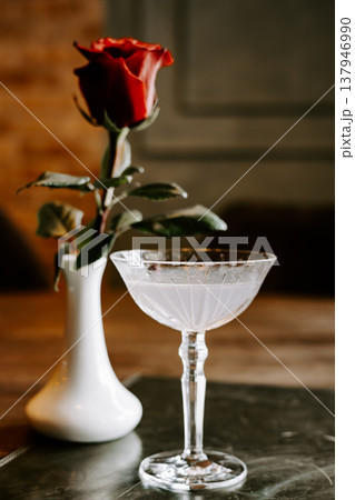 Top view of a crystal cocktail glass and a red rose in a white vase on a dark wooden table. Moody still life for romantic and celebration designs. Top view of a crystal cocktail glass and a red rose in a white vase on a dark wooden table. Moody still life for romantic and celebration designs. 137946990
