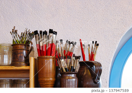 A wooden shelf in rustic artist studio or creative workshop, with glass jars and clay jugs filled with paintbrushes A wooden shelf in rustic artist studio or creative workshop, with glass jars and clay jugs filled with paintbrushes 137947589