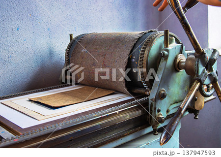 Closeup of a hand operated manual printing press with a layered printmaking plate and paper running through the rollers. 137947593