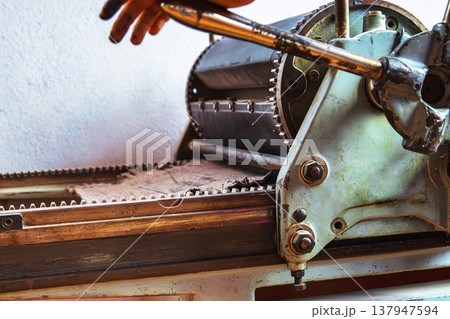 A detailed closeup of a traditional manual printing press with visible roller gears and crank handle, partially operated by hand. A detailed closeup of a traditional manual printing press with visible roller gears and crank handle, partially operated by hand. 137947594