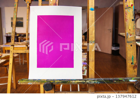 A single magenta monoprint is centered on a white sheet of paper, standing upright on a well used wooden easel in an art classroom. A single magenta monoprint is centered on a white sheet of paper, standing upright on a well used wooden easel in an art classroom. 137947612
