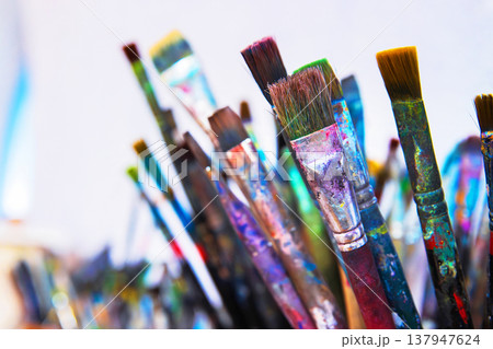 A cluster of well used paintbrushes with colorful dried paint on their handles and bristles in container in creative art studio. A cluster of well used paintbrushes with colorful dried paint on their handles and bristles in container in creative art studio. 137947624