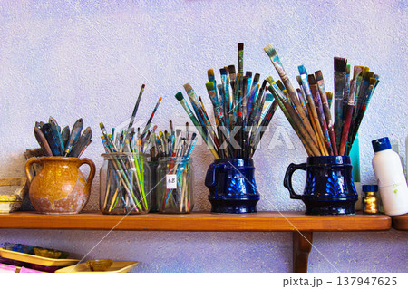 Various paintbrushes of different shapes and size, arranged in ceramic mugs and glass jars on a wooden shelf against a textured wall. Various paintbrushes of different shapes and size, arranged in ceramic mugs and glass jars on a wooden shelf against a textured wall. 137947625