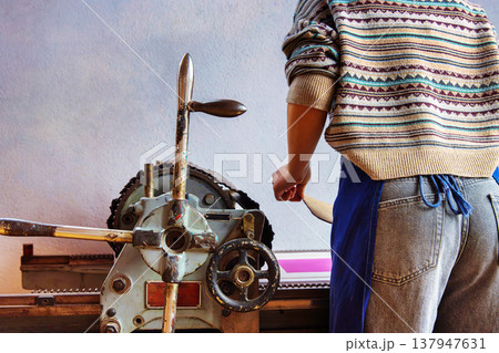 A printmaker in a patterned sweater and apron operates a traditional mechanical printing press. Back view of the artist during the printmaking process A printmaker in a patterned sweater and apron operates a traditional mechanical printing press. Back view of the artist during the printmaking process 137947631