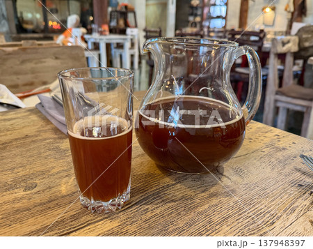 Glass jug and tall glass with kvass stand on wooden table in restaurant interior, traditional fermented drink with dark amber color, light foam. Beverage serving, drink presentation, restaurant menu Glass jug and tall glass with kvass stand on wooden table in restaurant interior, traditional fermented drink with dark amber color, light foam. Beverage serving, drink presentation, restaurant menu 137948397