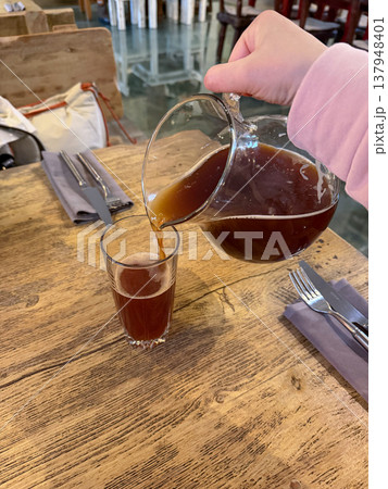Hand pours kvass from glass jug into tall glass on wooden table in restaurant setting, traditional fermented drink with dark color and foam. Beverage serving, drink presentation, restaurant menu Hand pours kvass from glass jug into tall glass on wooden table in restaurant setting, traditional fermented drink with dark color and foam. Beverage serving, drink presentation, restaurant menu 137948401