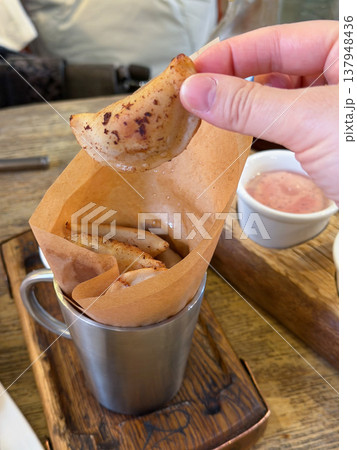 Hand holds small fried dumpling above metal cup with paper liner and sauce on wooden table in restaurant setting, crispy snack with golden texture. Food serving, street food menu, snack Hand holds small fried dumpling above metal cup with paper liner and sauce on wooden table in restaurant setting, crispy snack with golden texture. Food serving, street food menu, snack 137948436