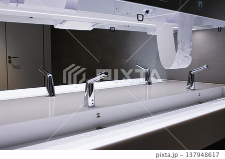 Interior of a modern public restroom featuring a long white sink with multiple chrome automatic faucets, mirror and a soap and towel dispensers. Interior of a modern public restroom featuring a long white sink with multiple chrome automatic faucets, mirror and a soap and towel dispensers. 137948617