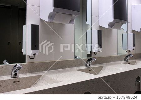 Interior of a modern public restroom with multiple sinks with chrome automatic faucets, mirrors, soap and towel dispensers. Interior of a modern public restroom with multiple sinks with chrome automatic faucets, mirrors, soap and towel dispensers. 137948624