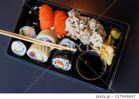 A takeaway sushi meal in a black plastic tray with salmon nigiri, maki rolls, ginger, wasabi, and soy sauce with chopsticks over the tray. Selective focus A takeaway sushi meal in a black plastic tray with salmon nigiri, maki rolls, ginger, wasabi, and soy sauce with chopsticks over the tray. Selective focus 137948647