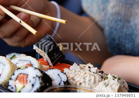 Closeup of the person hand picking up a sushi roll with chopsticks over a black tray in a modern cafe. Selective focus 137948651