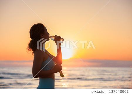 Fit woman drinking water at the beach after running outdoors in the morning. Sporty female relaxing after training. 137948961