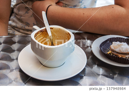Finished espresso cup, spoon and powdered italian pastry on metal cafe table. Young woman arm in the background. Selective focus Finished espresso cup, spoon and powdered italian pastry on metal cafe table. Young woman arm in the background. Selective focus 137948984