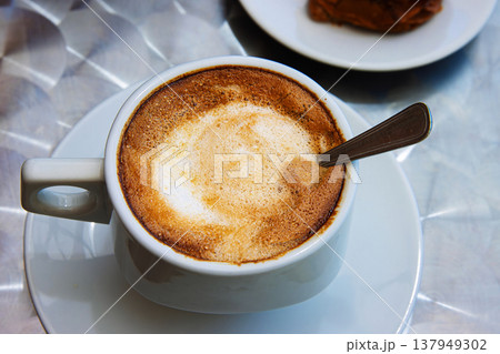 Top view of a cappuccino with creamy milk foam in a round white ceramic cup on a saucer served on a metallic table in outdoor cafe. Selective focus. Top view of a cappuccino with creamy milk foam in a round white ceramic cup on a saucer served on a metallic table in outdoor cafe. Selective focus. 137949302