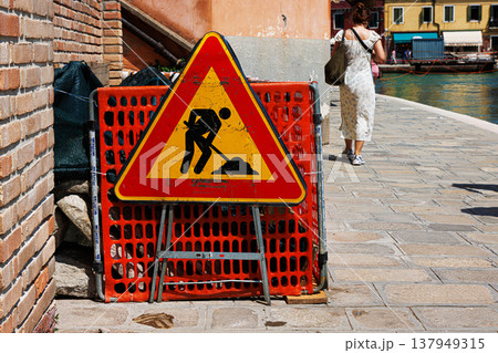 Murano, Province of Venice, Italy, June 14th, 2025. Closeup of a road work warning sign in the construction zone on a city sidewalk with an orange safety barrier and blurred pedestrian Murano, Province of Venice, Italy, June 14th, 2025. Closeup of a road work warning sign in the construction zone on a city sidewalk with an orange safety barrier and blurred pedestrian 137949315