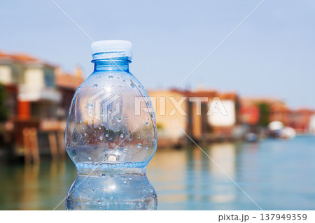 A half full plastic bottle of sparkling mineral water rests on a bridge railing in Murano, Italy. Blue sky, canal with boats and colorful buildings on hot summer day. A half full plastic bottle of sparkling mineral water rests on a bridge railing in Murano, Italy. Blue sky, canal with boats and colorful buildings on hot summer day. 137949359