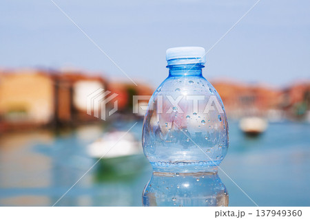 A half full plastic bottle of sparkling mineral water rests on a bridge railing in Murano, Italy. Blue sky, canal with boats and colorful buildings on hot summer day. 137949360