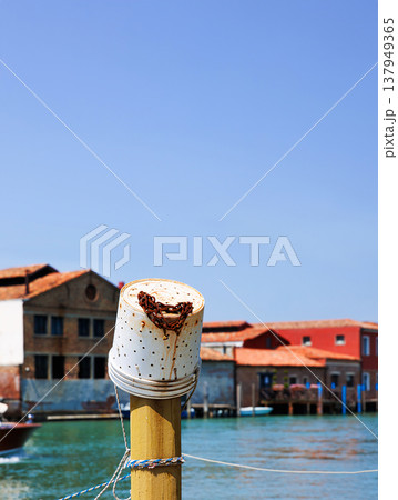 Mooring post with plastic fishing bucket with rusty chain and a motorboat moving along the turquoise canal water passing  colorful buildings in Murano, Venetian Lagoon 137949365