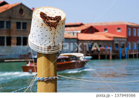 Mooring post with plastic fishing bucket with rusty chain and a motorboat moving along the turquoise canal water passing colorful buildings in Murano, Venetian Lagoon Mooring post with plastic fishing bucket with rusty chain and a motorboat moving along the turquoise canal water passing colorful buildings in Murano, Venetian Lagoon 137949366