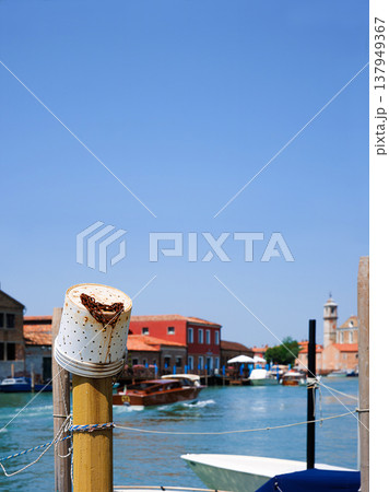 Murano, Province of Venice, Italy, June 14th, 2025. Mooring post with plastic fishing bucket with rusty chain and a motorboat moving along the turquoise canal water passing colorful buildings. Murano, Province of Venice, Italy, June 14th, 2025. Mooring post with plastic fishing bucket with rusty chain and a motorboat moving along the turquoise canal water passing colorful buildings. 137949367