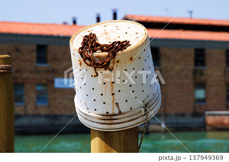 A reused plastic bucket with holes, transformed into a fishing container, hangs on an old wooden mooring post known as a palina Canal in Murano, Venetian Lagoon 137949369