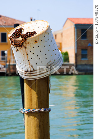 A reused plastic bucket with holes, transformed into a fishing container, hangs on an old wooden mooring post known as a palina Canal in Murano, Venetian Lagoon A reused plastic bucket with holes, transformed into a fishing container, hangs on an old wooden mooring post known as a palina Canal in Murano, Venetian Lagoon 137949370