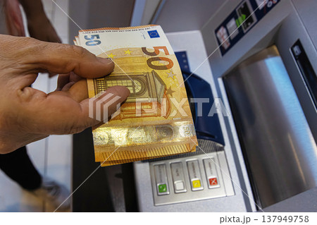 Closeup of a person holding multiple 50 euro banknotes in front of an ATM keypad. The terminal features colored confirmation buttons and a money slot. 137949758
