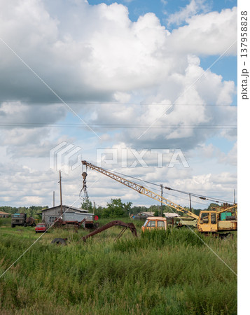 Old yellow construction crane standing on grassy field near rural building. Abandoned heavy machinery at industrial site. Rusty steel equipment in countryside under cloudy blue sky. Old yellow construction crane standing on grassy field near rural building. Abandoned heavy machinery at industrial site. Rusty steel equipment in countryside under cloudy blue sky. 137958828