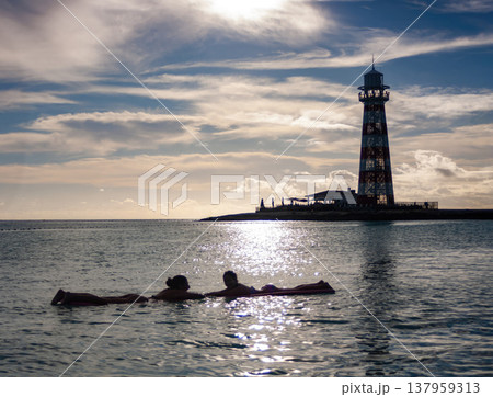 Silhouette of two young people resting on the water in a summer cloudy afternoon with a lighthouse in the background. Silhouette of two young people resting on the water in a summer cloudy afternoon with a lighthouse in the background. 137959313