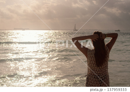 Woman standing in the ocean water at sunset observing the coastal view and sailboat, embodying summer relaxation and a serene vacation lifestyle Woman standing in the ocean water at sunset observing the coastal view and sailboat, embodying summer relaxation and a serene vacation lifestyle 137959333
