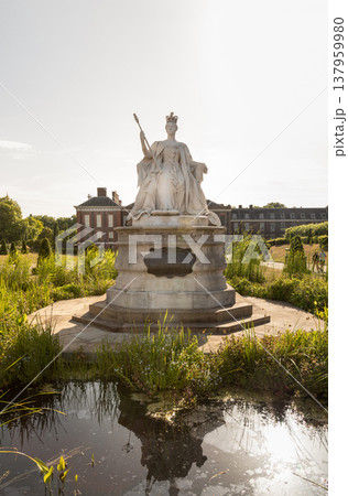Captured from a low angle, a white marble statue of the young Queen Victoria rises against the soft glow of morning light, projecting an air of quiet majesty. The tranquil pond in the foreground Captured from a low angle, a white marble statue of the young Queen Victoria rises against the soft glow of morning light, projecting an air of quiet majesty. The tranquil pond in the foreground 137959980