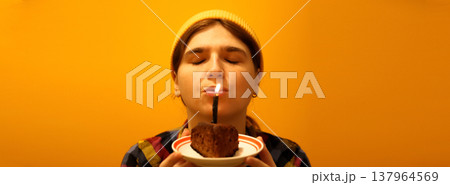 Young woman with closed eyes blowing out a candle on a chocolate cake. Birthday celebration and making a wish concept on a yellow background. Happy Birthday banner Young woman with closed eyes blowing out a candle on a chocolate cake. Birthday celebration and making a wish concept on a yellow background. Happy Birthday banner 137964569