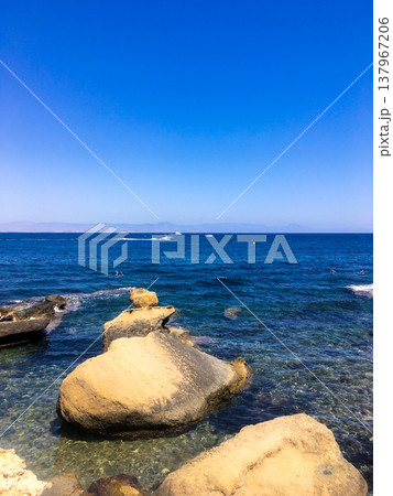 Clear blue sea and large rocks along coastline of Tabarca island at Spain. Gentle waves moving towards shore, boats pass by on horizon under bright blue sky. Tourism and holiday 137967206