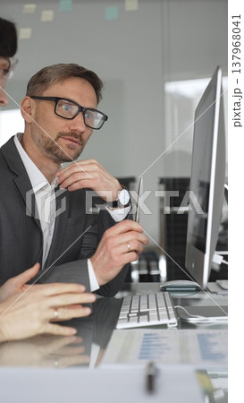 Business professional analyzing growing financial data on computer monitor with colleague, pointing at screen while discussing market trends and economy strategy in modern office, vertical view 137968041