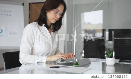 Businesswoman in white blouse and glasses sitting at a modern desk, working with a tablet in a contemporary office, focusing on digital tasks, using technology for efficient workflow Businesswoman in white blouse and glasses sitting at a modern desk, working with a tablet in a contemporary office, focusing on digital tasks, using technology for efficient workflow 137968133