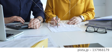 Two accountants collaborating in a well lit office, analyzing financial data and reviewing statements with a calculator and pen. Audit and taxes theme in business Two accountants collaborating in a well lit office, analyzing financial data and reviewing statements with a calculator and pen. Audit and taxes theme in business 137968179