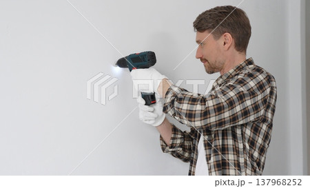 Man construction worker in checked shirt and safety gloves is screwing a screw into the wall using a cordless screwdriver, carrying out a careful home renovation. Horizontal portrait view Man construction worker in checked shirt and safety gloves is screwing a screw into the wall using a cordless screwdriver, carrying out a careful home renovation. Horizontal portrait view 137968252