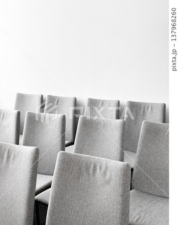 Gray chairs in straight row against plain white wall. Fabric with minimalist design, room for meetings or group gatherings. Clean and organized environment for discussions or presentations Gray chairs in straight row against plain white wall. Fabric with minimalist design, room for meetings or group gatherings. Clean and organized environment for discussions or presentations 137968260