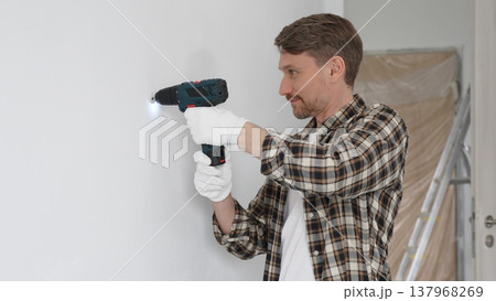 Man construction worker in checked shirt and safety gloves is screwing a screw into the wall using a cordless screwdriver, carrying out a careful home renovation. Horizontal portrait view 137968269