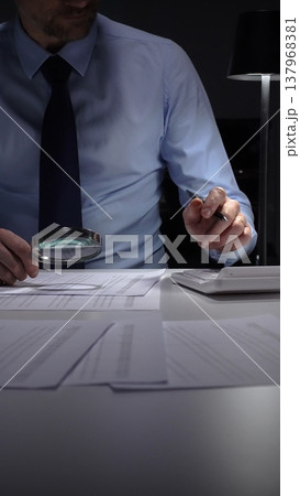 Businessman diligently auditing financial documents with a magnifying glass under focused lamp light. Audit and taxes in business 137968381