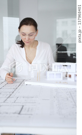 Female architect in eyeglasses concentrating on a detailed building model and blueprints, carefully planning a modern architectural design in a professional office 137968401