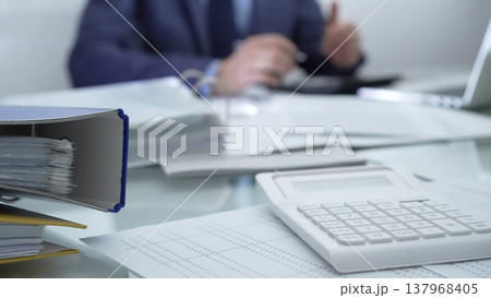 Calculator, pen, laptop and documents are lying on accountant's desk. Businessman wearing blue suit is working with documents on the background. Business people, audit concept Calculator, pen, laptop and documents are lying on accountant's desk. Businessman wearing blue suit is working with documents on the background. Business people, audit concept 137968405