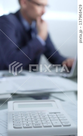 Calculator, pen, laptop and documents are lying on accountant's desk. Businessman wearing blue suit is working with documents on the background. Vertical view. Business and audit concept Calculator, pen, laptop and documents are lying on accountant's desk. Businessman wearing blue suit is working with documents on the background. Vertical view. Business and audit concept 137968429