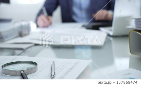 Magnifying glass, calculator, pen, laptop and documents are lying on accountant's desk. Businessman wearing blue suit is working with documents on the background. Business people and audit concept 137968474