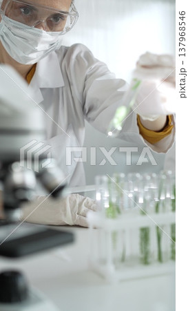 Woman scientist wearing lab coat, white gloves, face mask and protective glasses, is holding a test tube with plants inside near microscope in laboratory, vertical portrait view. Science and medicine Woman scientist wearing lab coat, white gloves, face mask and protective glasses, is holding a test tube with plants inside near microscope in laboratory, vertical portrait view. Science and medicine 137968546