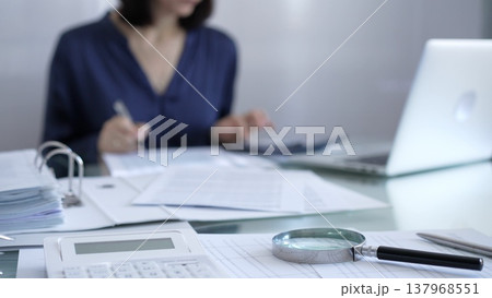Magnifying glass, white calculator, pen and documents are lying on accountant's desk. Businesswoman wearing blue dress is working with documents on the background. Business and audit concept Magnifying glass, white calculator, pen and documents are lying on accountant's desk. Businesswoman wearing blue dress is working with documents on the background. Business and audit concept 137968551