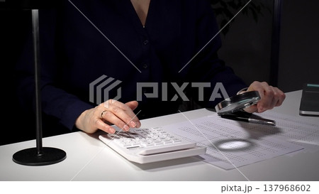 Person checking financial documents and numbers with a magnifying glass, analyzing business data for budgeting and taxes at night. Audit and taxes in business Person checking financial documents and numbers with a magnifying glass, analyzing business data for budgeting and taxes at night. Audit and taxes in business 137968602